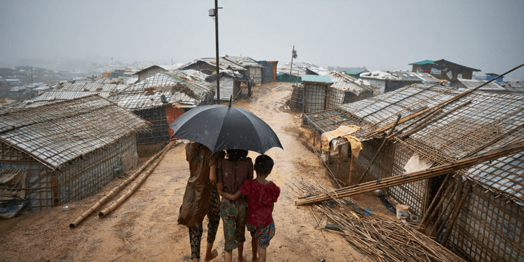 Three Rohingya children huddle under an umbrella during a downpour in Cox's Bazar, Bangladesh