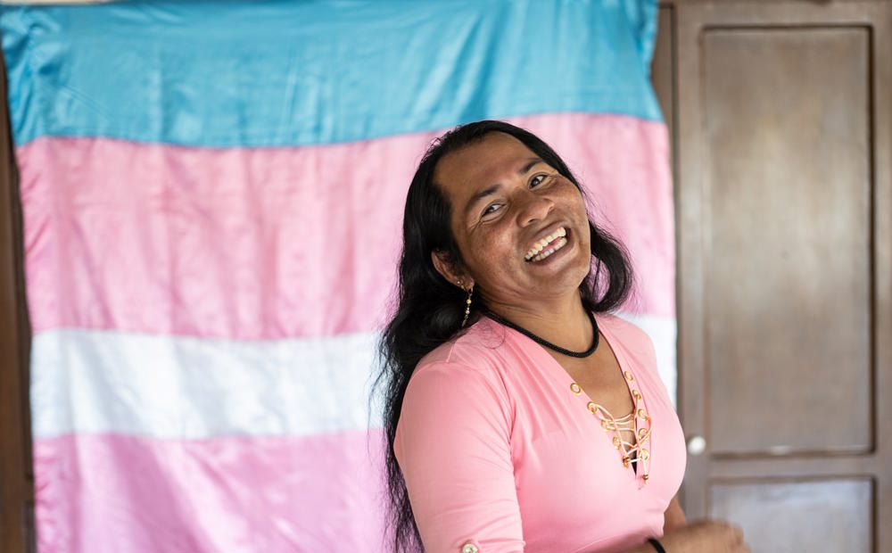 Synthia, an indigenous trans woman from Colombia, smiles at the camera, in front of the transgender flag