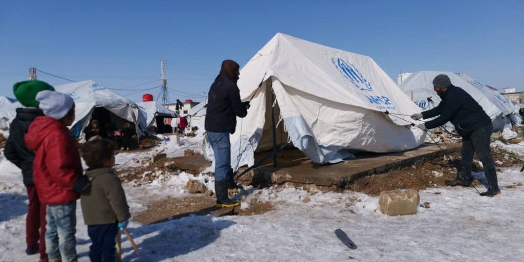 Two men are repairing a UNHCR tent. They are surrounded by snow and refugee children are looking at them. UNHCR with its partners distributs blankets, mattresses, clothing and reinstals tents to families affected by the snow storm in the camps of north-east Syria ; More than 13 million Syrians have been displaced in the past 10 years.