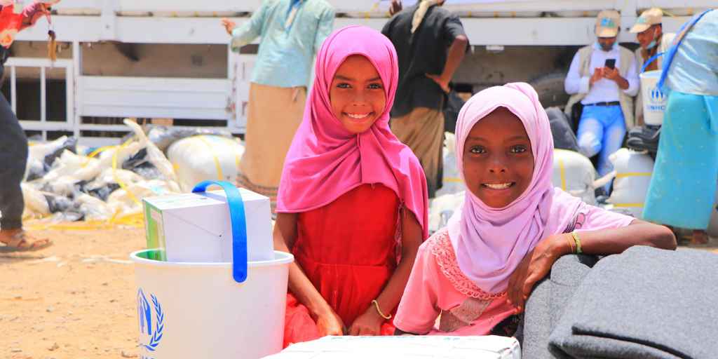 Two refugee girls smile surrounded by UNHCR supplies