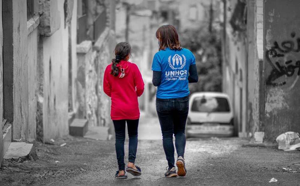 UNHCR staff and a refugee girl walking together. The background, a street seemed to be torn by war and disaster, is black and white