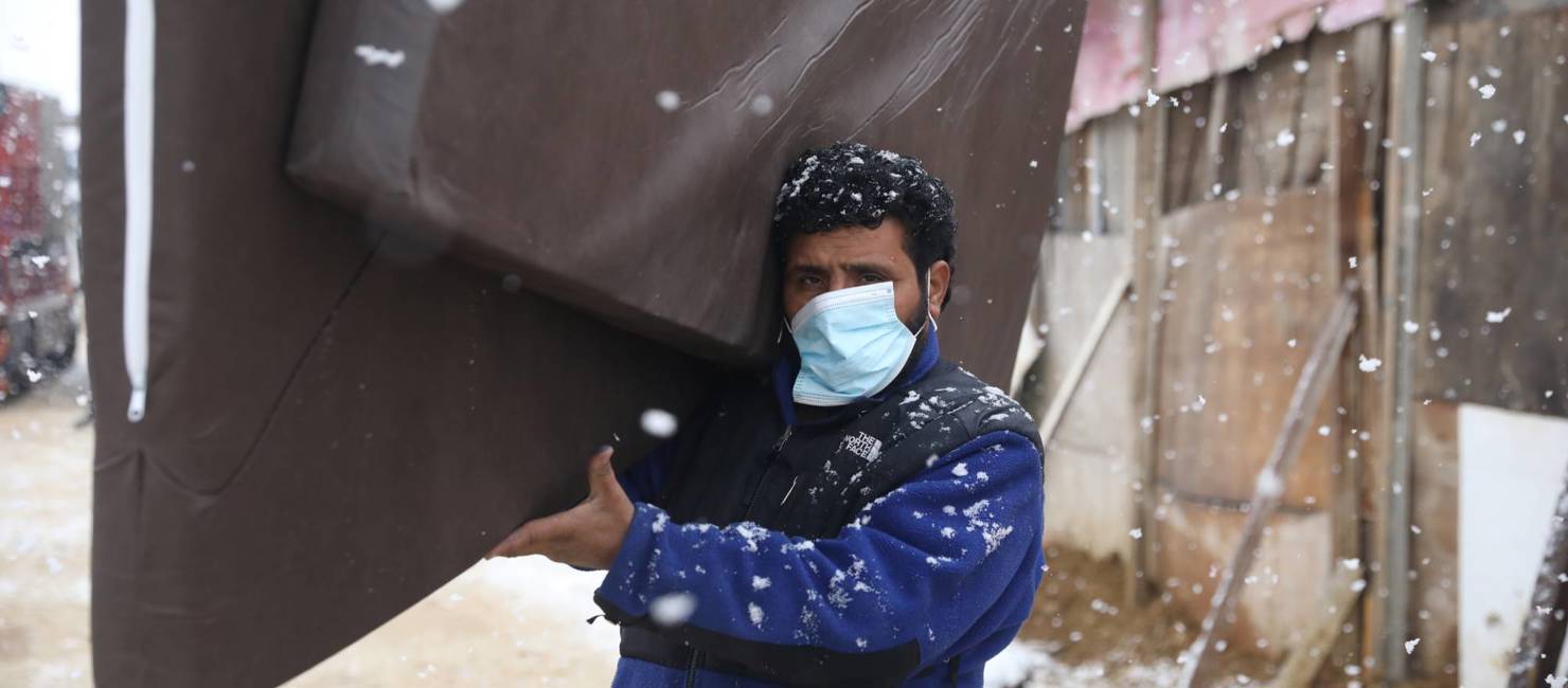 A refugee carries mattresses through a makeshift settlement while snow falls.