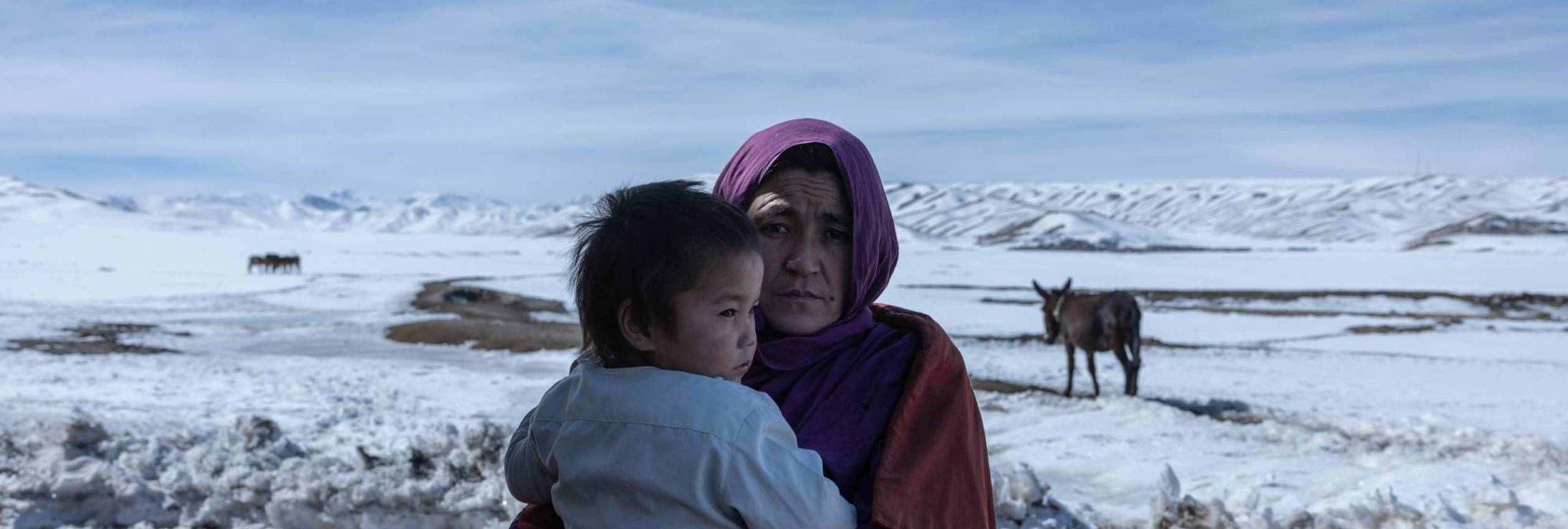 Afghan woman Zahra*, 34, looks at the camera while holding her baby daughter, a snowy landscape in the background.