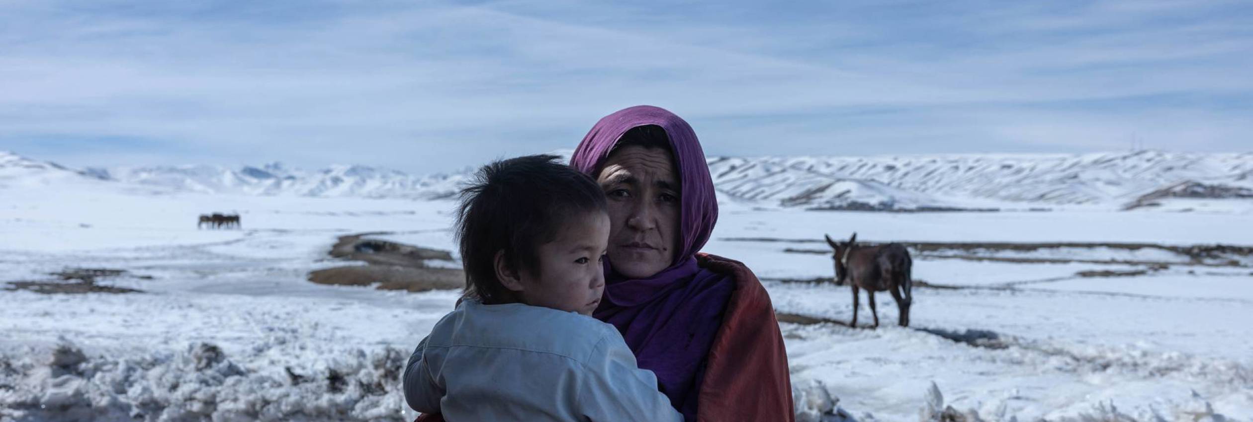 Afghan woman Zahra*, 34, looks at the camera while holding her baby daughter, a snowy landscape in the background.