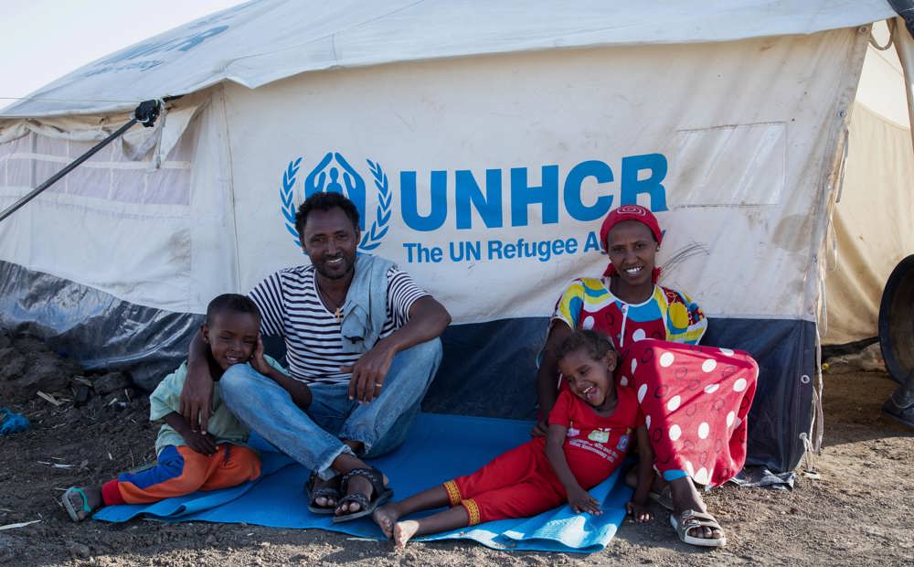 Family of Ethiopian refugees smiling. UNHCR tent in the background. Ethiopian refugees Tsiruy Dagnew, 40, Abeba Kalayu, 32, with their children Semere, 5, and Eleni, 3, enjoy the sun near their tent at Tunaydbah camp in Sudan. Many of its refugees have found innovative and resourceful ways to begin rebuilding their lives. 