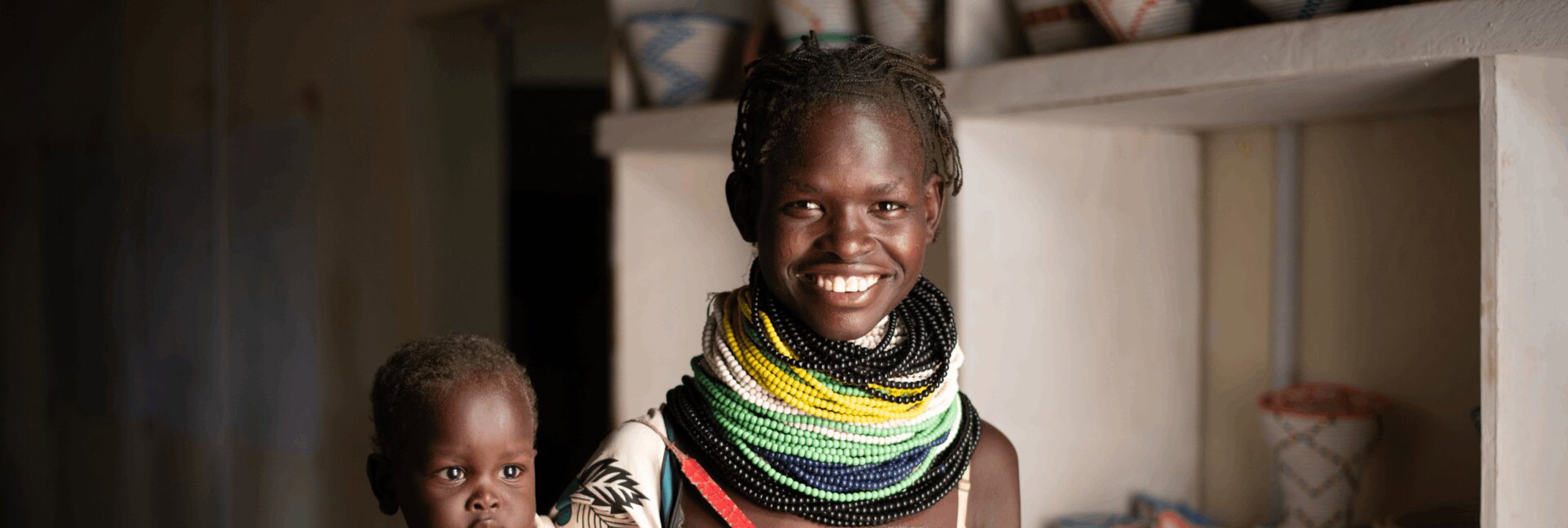 Lucy Akai, a Kenya woman, holds her baby and smiles at the camera. She is wearing brightly coloured beaded jewelery.