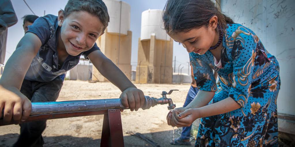 A boy and a girl in a refugee camp in Syria. The girl is having some water from the communal water tap. Providing potable water and raising awareness among children about the importance of hand washing is essential for preventing the spread of COVID-19.