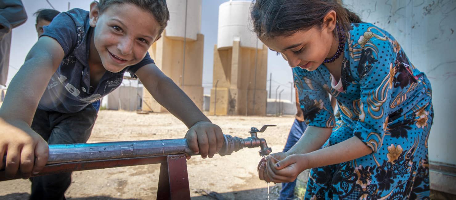 A boy and a girl in a refugee camp in Syria. The girl is having some water from the communal water tap. Providing potable water and raising awareness among children about the importance of hand washing is essential for preventing the spread of COVID-19.