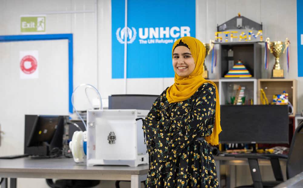 Woman smiles at camera with UNHCR sign behind her. UNHCR’s Innovation Lab promotes STEM for refugee girls. 