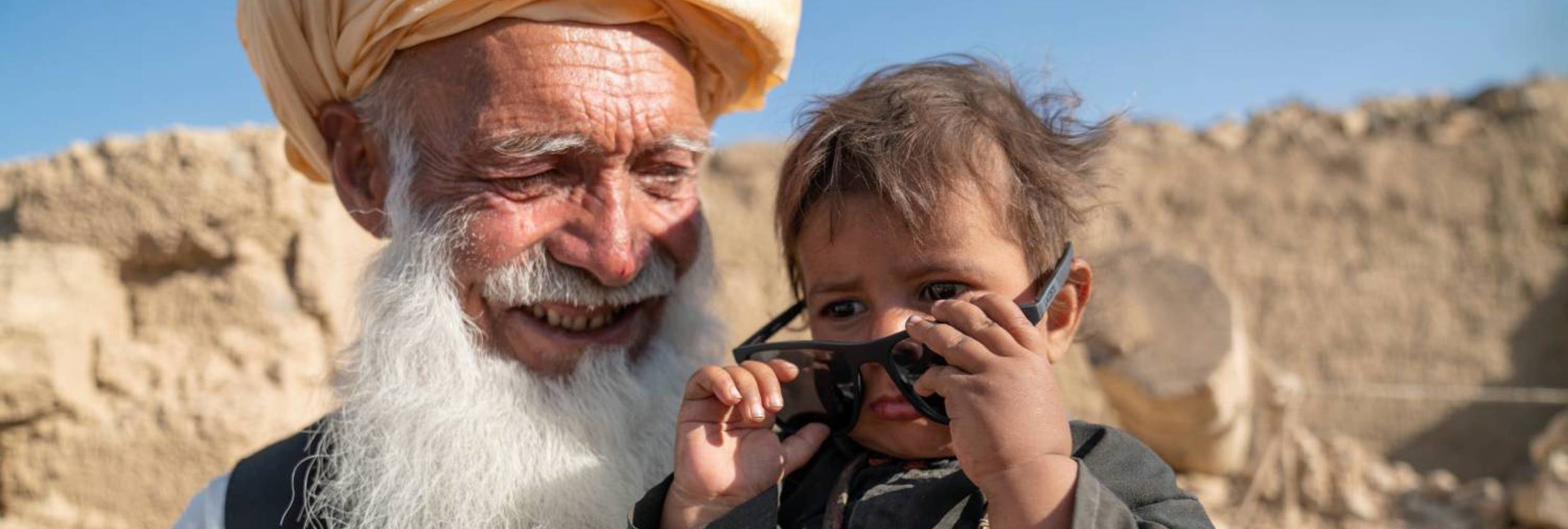 A boy puts on sunglasses while his grandfather holds him, smiling. In Afghanistan, UNHCR is providing earthquake-resilient houses for displaced families.